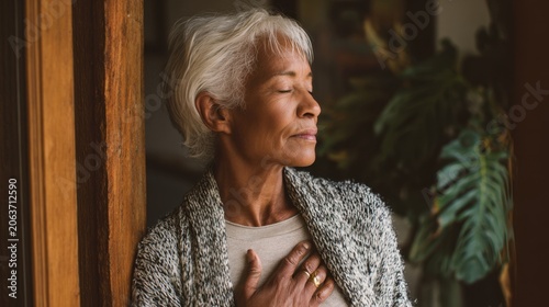 Senior woman with closed eyes and a serene expression standing indoors with her hand on her chest, practicing mindfulness and deep breathing