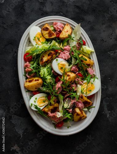 Salad with canned tuna, vegetables, arugula, baked potatoes and boiled eggs on a dark background, top view