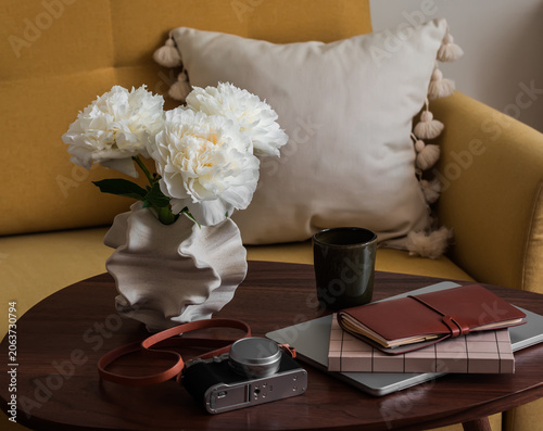 A laptop, a vase with peonies, a camera, a cup of coffee on a wooden table next to a yellow sofa