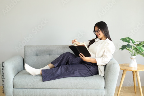 A young woman sits relaxed on the sofa at home, reading a book with a smile.