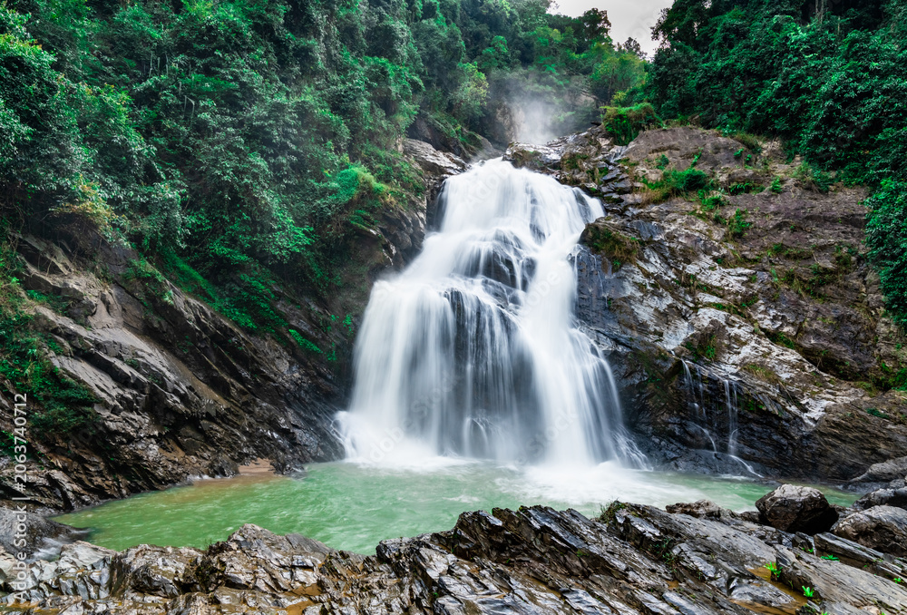 Obraz premium Beautiful waterfall at the mountain with blue sky and white cumulus clouds. Waterfall in tropical green tree forest. Waterfall is flowing in jungle. Nature abstract background. Granite rock mountain.