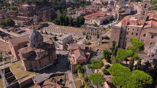Cinematic aerial view in Rome Italy showcasing Roman Forum with ancient temple columns, triumphal arches, historic church domes, terracotta rooftops, and lush green trees under bright summer sunlight.