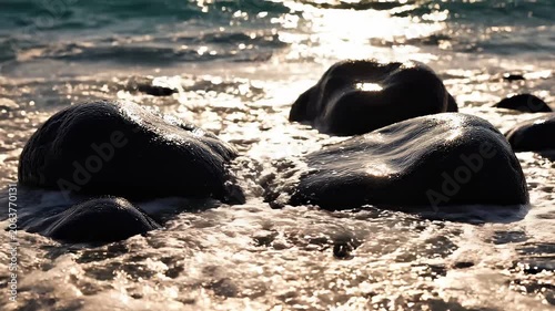 Close-up shot of wet rocks in the gentle surf of the ocean with shimmering sunlight reflecting on the water's surface