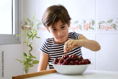 Young boy in striped shirt reaching into bowl for cherry in sunlit kitchen setting