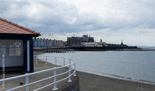 A painted wooden seaside shelter with benches on a coastal promenade overlooking calm ocean waters beneath a dramatic cloudy sky, evoking quiet seaside charm