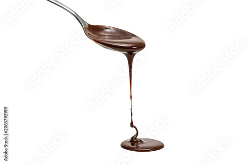 A silver metal spoon pouring thick liquid chocolate syrup that forms a small pool at the bottom, captured against a solid