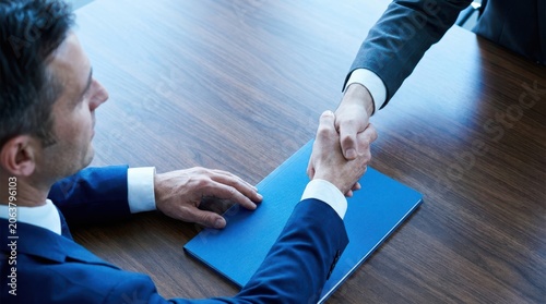 Two professional business partners shaking hands over blue folder on office wooden table during corporate agreement.