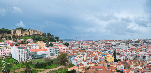 Lisbon panorama showing the historic Sao Jorge Castle dominating the skyline, with traditional buildings spread across hillsides and a suspension bridge crossing the Tagus river under a cloudy sky