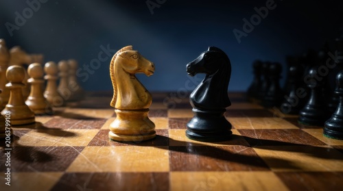 Chess game strategy concept with white and black knight pieces facing each other on wooden board under dramatic light.
