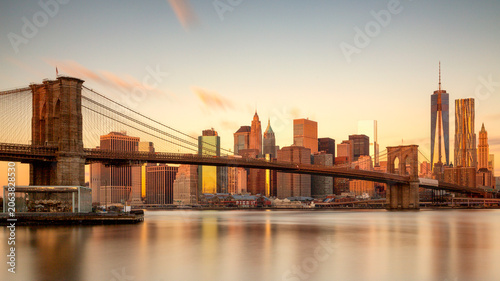 Brooklyn Bridge and the iconic Lower Manhattan skyline glow during a golden hour sunset over the East River, capturing the essence of urban tourism in New York City.