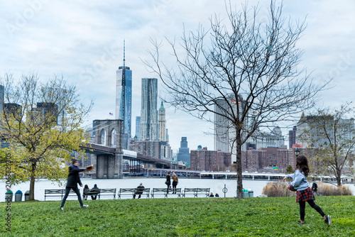 People playing catch on a green park lawn, embodying an active outdoor lifestyle with the Brooklyn Bridge and Lower Manhattan skyline background, New York City, USA.
