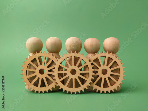 Five blank wooden peg dolls stand lined up behind three interconnected decorative wooden cogs on green background