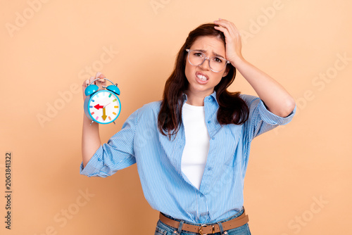 Принти на полотні Stressed young woman holding a blue alarm clock with anxious expression, worried