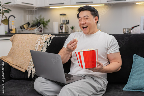 Konstfotografi Asian man at home streams comedy on laptop holds bowl of popcorn laughs hard and smiles wide feels joy