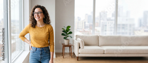 A beautiful young woman with curly hair and glasses standing happily near a window in a bright modern apartment with a cozy sofa