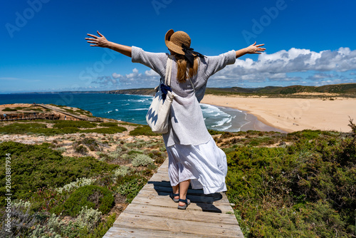 Beautiful middle-aged woman tourist walking down wooden stairs to sandy Bordeira beach and enjoying vacations in Algarve Portugal on summer day. Back view
