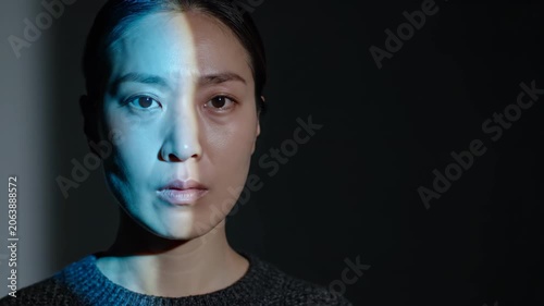Asian woman face illuminated by dual split lighting. Half of face lit by blue light while other half is in shadow. Concept of duality and contrast in portrait