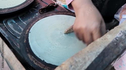 A batter is spread thinly using a wooden tool to form a crepe. 