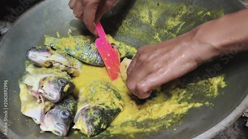 A person is marinating several whole fish with a yellow spice paste in a large metal wok. 