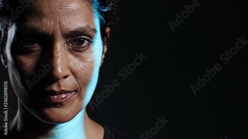 Mature Indian woman face with dramatic blue light on dark background. Close up portrait of female expressive gaze under studio lighting transition for media content