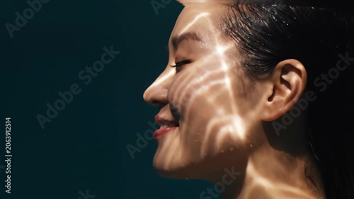 Asian woman submerged in water with dynamic light refraction on skin. Side view close-up of face enjoying water therapy. Skin care and wellness concept in motion