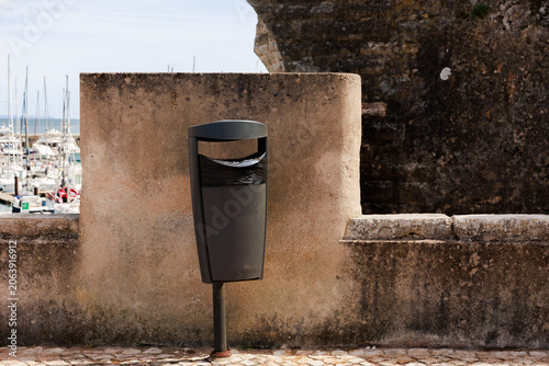 Cuadro en lienzo Dark grey public trash receptacle mounted on a pole against a textured stone wall overlooking a marina with boats
