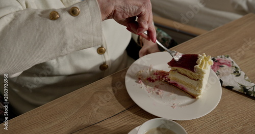 Older woman savoring delicious layered cake. Close-up shot of hand holding fork. Creamy dessert with berry topping on plate. Cozy and relaxed atmosphere.