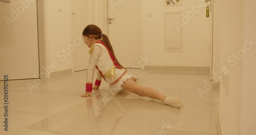 Young girl in costume practicing dance splits on shiny tile floor in white room, focused expression, elegant posture, determination and concentration