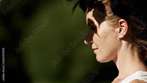 Caucasian woman in profile standing outdoors in sunny garden with tree leaf shadow on face. Daylight natural portrait series showing facial expression transition