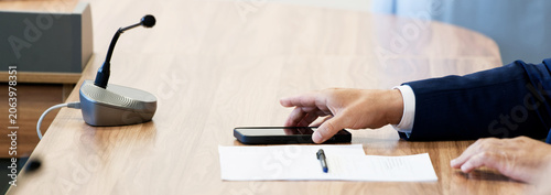 Hands of a politician or executive using a mobile phone during a formal boardroom meeting. Professional corporate environment with a desktop microphone and paperwork. Photo