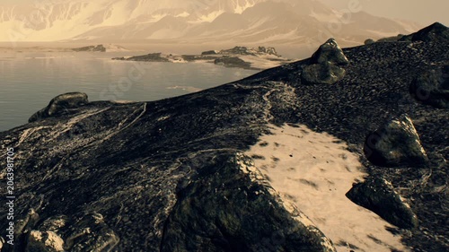 A rocky shore with water and mountains in the background. The sun is low, creating a warm light over the area. The landscape appears rugged and wild.