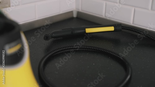 Close up shot of a yellow and black steam cleaner nozzle resting on a black speckled kitchen countertop with white subway tile backsplash visible in the background