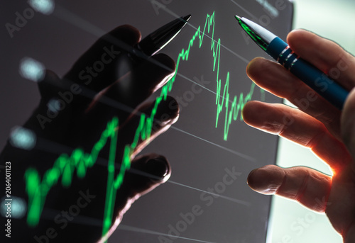 Hands pointing at a stock market graph on a screen with green line chart. 