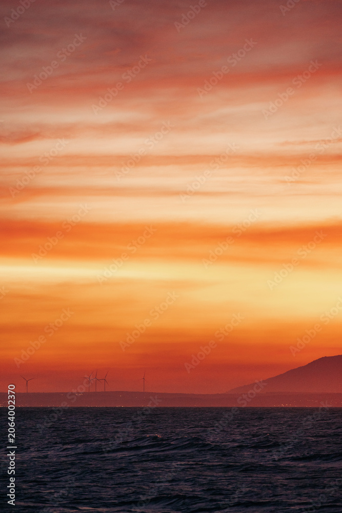 Fototapeta premium Stunning sunset over the ocean with distant wind turbines and hills on the horizon. Mui Ne, Vietnam