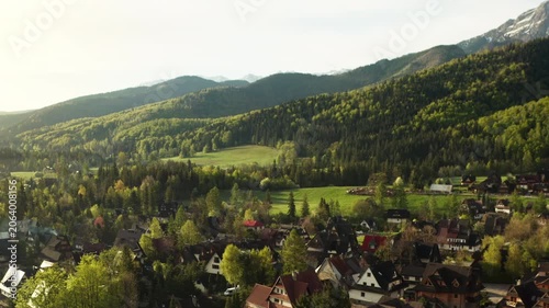 Traditional wooden houses and green meadows in mountain village at sunrise. Scenic alpine landscape with pine forest and Tatra peaks near Zakopane resort in Poland during spring morning.