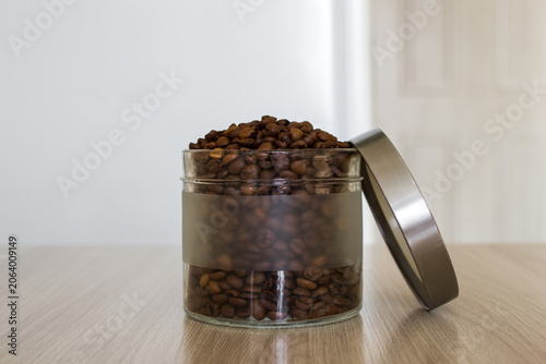 Open glass jar with coffee beans on wooden table in light kitchen.