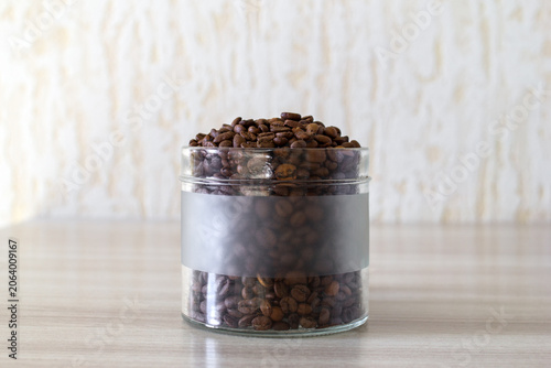Open glass jar full of aromatic coffee beans on wooden table in light kitchen.