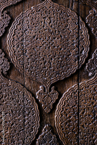 Ornate carved wooden door detail from Muhammad Amin Khan Madrasah in Khiva. Carved wood texture, pattern, background. Intricate floral arabesque medallion pattern on aged wood panels.