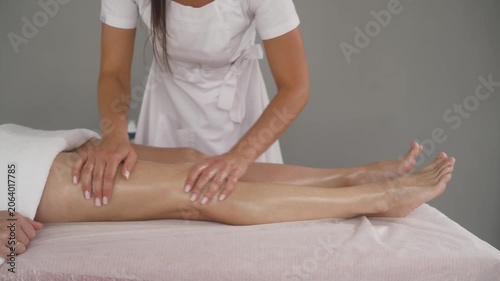 Professional masseuse in white uniform performs relaxing leg massage on client lying on pink table in spa salon. Therapist applies oil and strokes legs with hands
