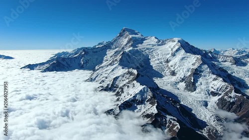 Aerial view of snow-covered mountain peaks and jagged ridges rising above a thick white sea of clouds and cloud inversion layer under a clear deep blue sky in high-altitude terrain.