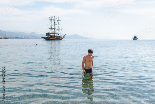 Young man walking in transparent Mediterranean sea water, Alanya, Turkey. Beautiful wooden sailing vessel in the background under soft sunlight. Horizontal medium shot, copy space in sky