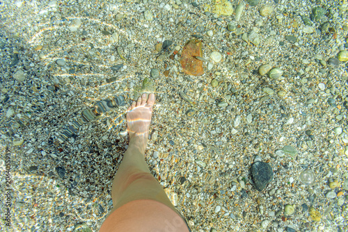 Close-up of small Mediterranean fish swimming near a human foot in clear turquoise water. Horizontal lifestyle shot from Alanya with sunlit pebble bottom and copy space for wellness design