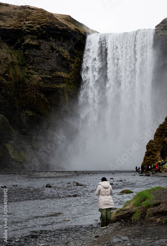 A person stands in front of Skogafoss waterfall in Iceland. The waterfall cascades down rocky cliffs while others explore the area. The sky is overcast and water splashes nearby.