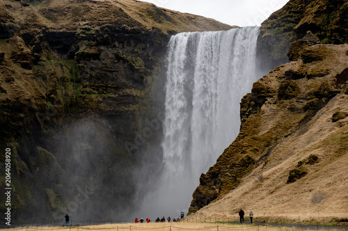 People walk along the pathway near Skogafoss waterfall in Iceland. Clouds cover the sky as visitors enjoy nature.