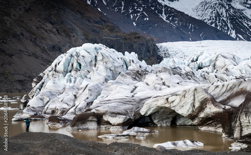 Drone aerial landscape of person in red jacket walking on a long, straight gravel road through vast, rugged icelandic highlands with snow capped mountains