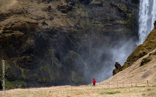 A side view of tourists in colorful jackets walking along a fenced grassy pathway at the base of the dramatic, rocky canyon cliffs of Skogafoss waterfall, with mist rising from the plunging water.