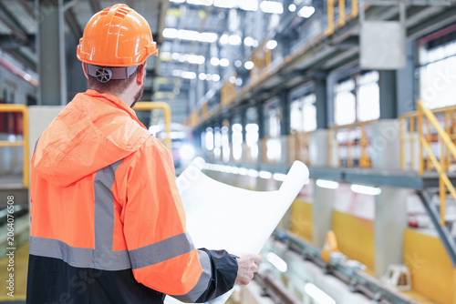 Architectural designer assesses intricate blueprints within brightly lit industrial warehouse featuring robust metal supports and advanced machinery equipment for detailed project evaluation.