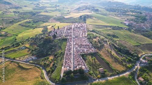 Aerial view of a historic cemetery with rows of tombs surrounded by rolling green hills and agricultural fields in Santa Caterina Villarmosa, Sicily, Italy.