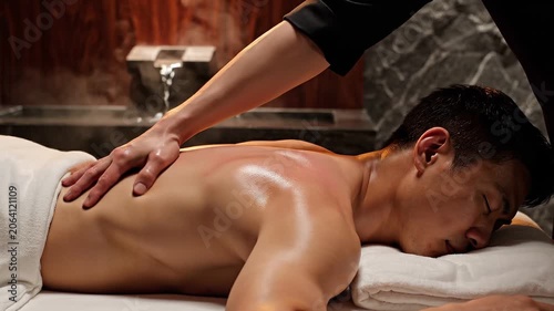 Masseur massaging back of asian man lying on massage table in spa center. Relaxation therapy session for body wellness and muscle health recovery in dark room.
