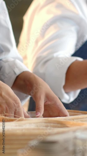 Vertical video: Order cueing hands in blue aprons lining bamboo steamer with wrappers at prep bench